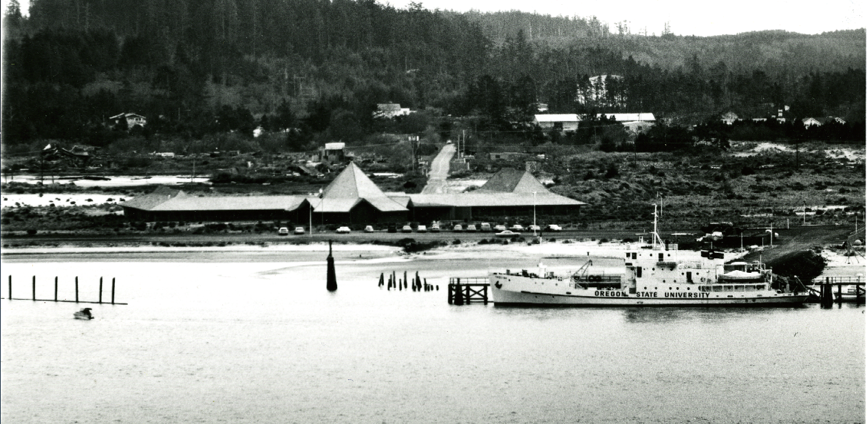 Black and white image of Marine Science Center from Yaquina Bay, facing South. Primary buildings shown, with forest in background. Research vessel Yaquina docked in front of center (foreground)
