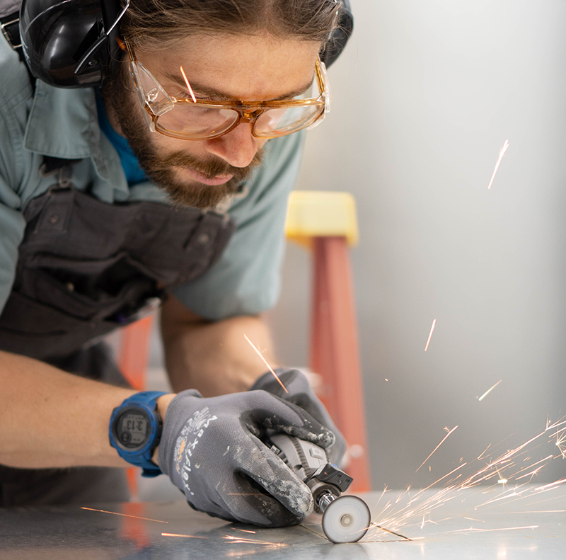 A man wearing saftey goggles and ear protection and work gloves uses a cutting tool on a metal sheet. Sparks are flying.