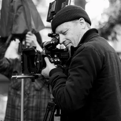 A black and white image of a man standing by a camera positioned on a tripod. 