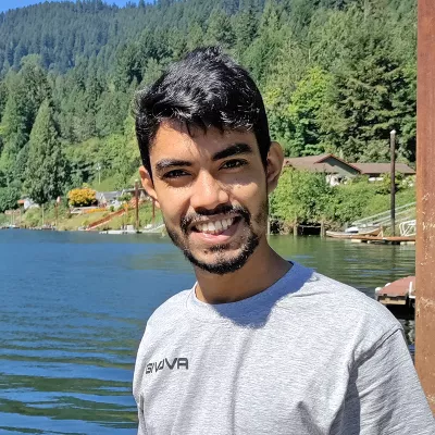 A young many with short black hair smiles into the camera. He is standing outside near a clear lake with a wooded hillside.
