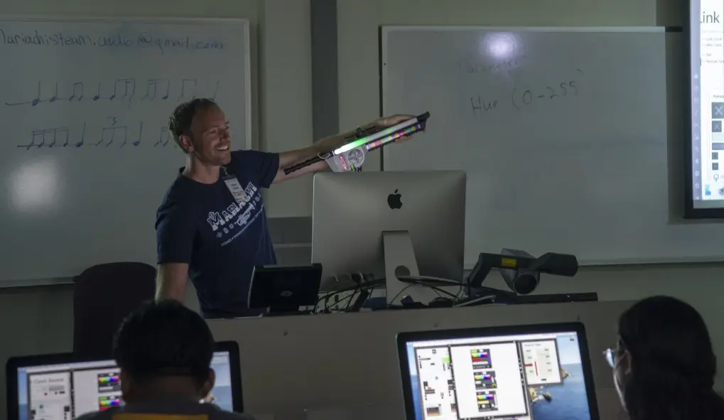 An instructor holds up an electronic musical instrument for a classroom of students to see.