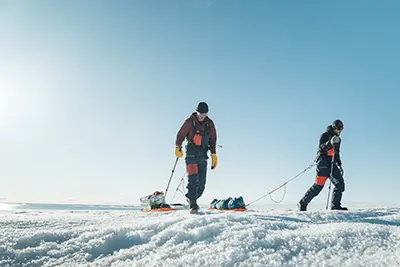 Two people pull sleds across a flat icy terrain.