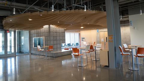 Lobby of the Gladys Valley Marine Science Building. A wooded wave sculpture hangs from the high ceilinig. Tall orange cafe chairs and tables are in lobby.