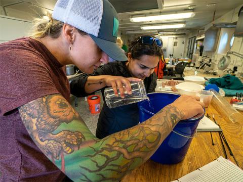 Two women with tattooed arms work to pour water in a bucket. 