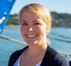 A woman with light blonde hair pulled back stands outside near water.